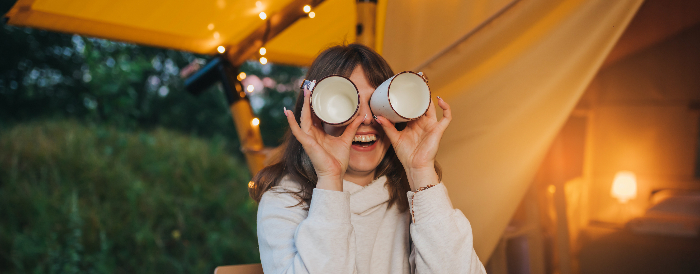 A woman smiling while covering her eyes with two mug cups.