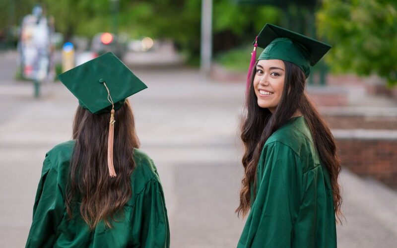 A young graduate wearing a green graduation gap and gown is looking back with a smile.