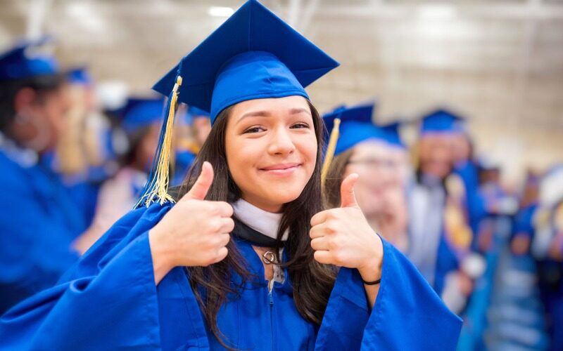 A young women wearing blue graduation cap and gown smiling at the camera with two thumbs up.