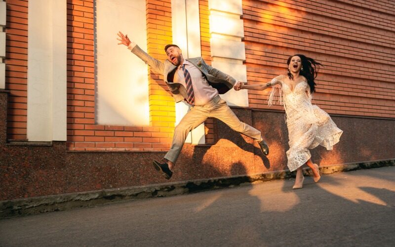 A bride and a groom holding hands and running on the street with funny expressions.